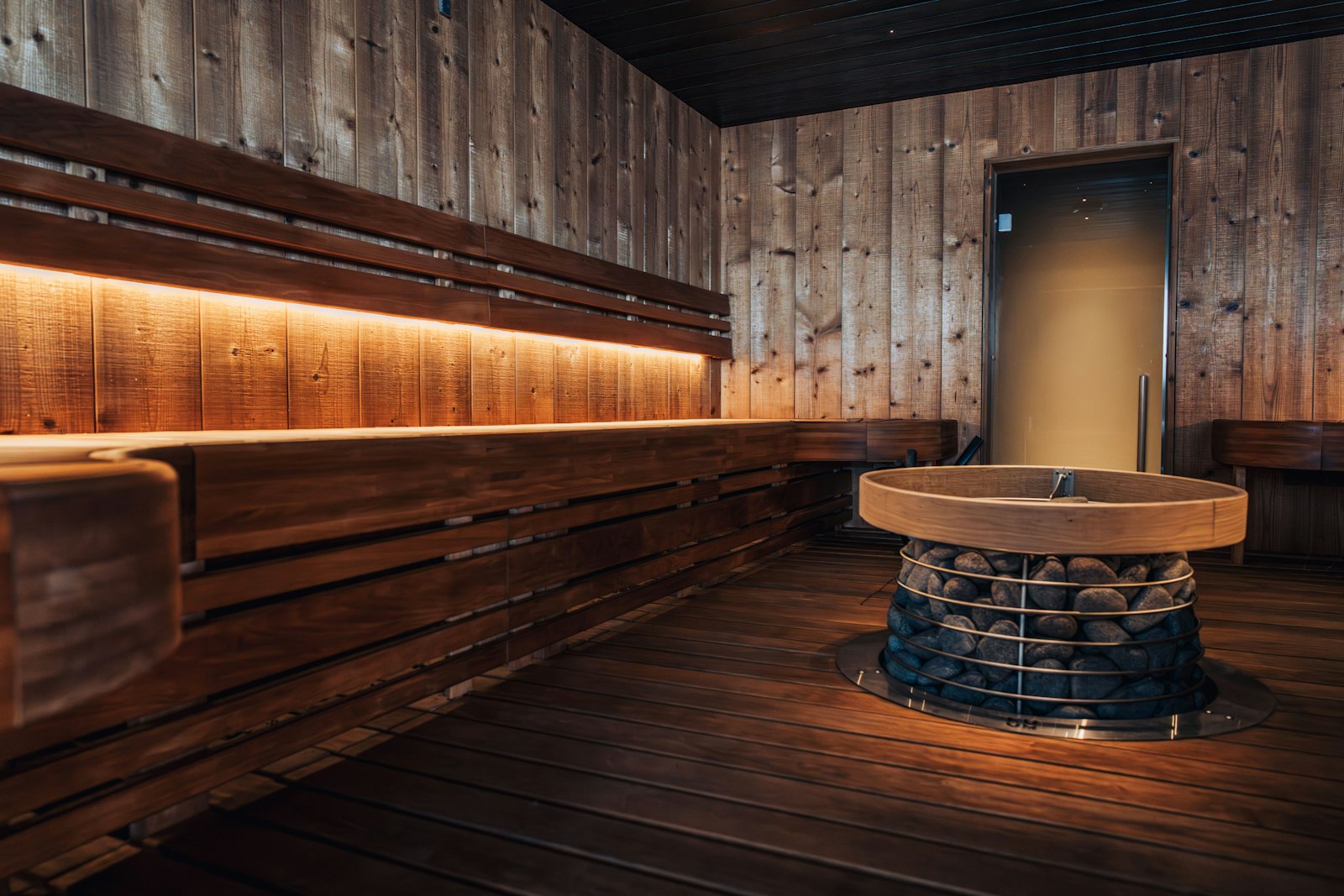 Sauna aus dunklem Holz mit langer Bank und rundem Steinofen