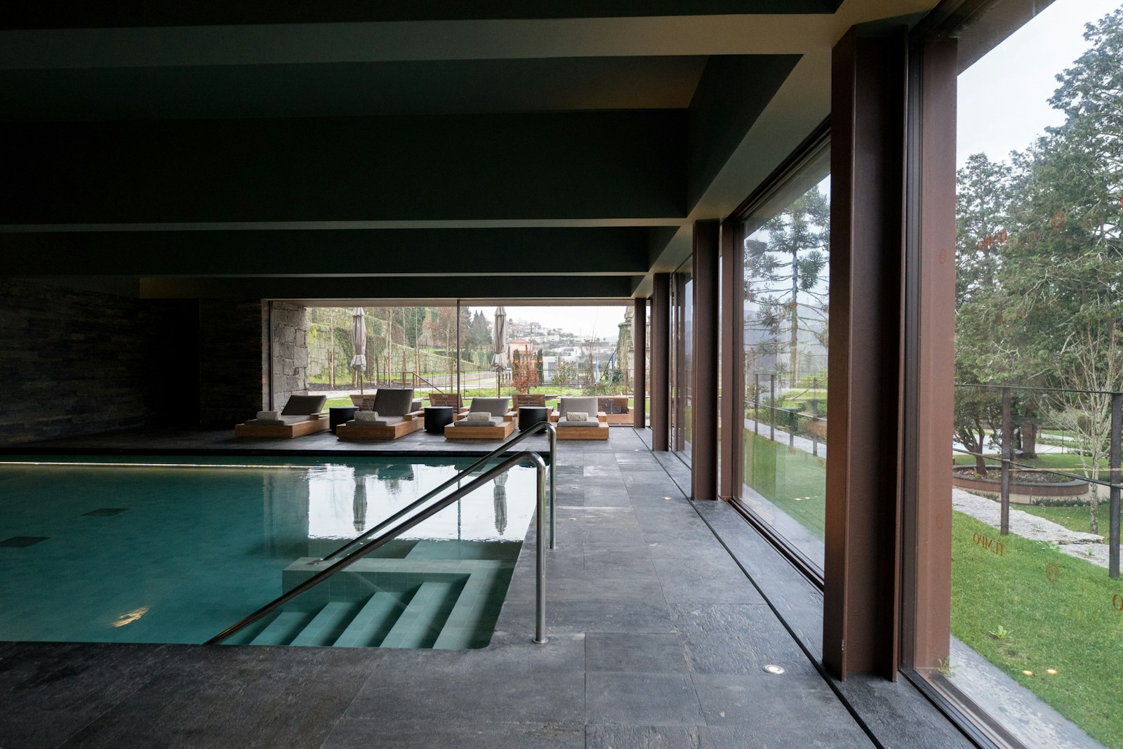 Innenpool mit dunklem Steinboden und raumhohen Fenstern, dahinter der Garten