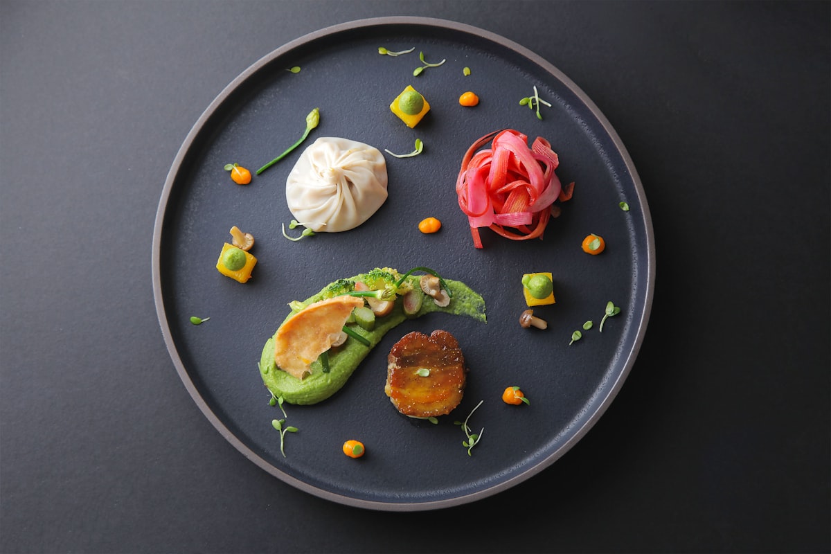 Dunkler Teller mit Erbsencreme, glasiertem Medaillon, hellem Teigbeutel und eingelegtem Rhabarber